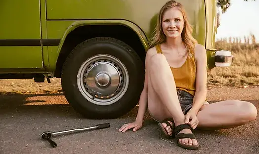 A woman sat in front of her camper van wheel which she has just changed. She looks happy.